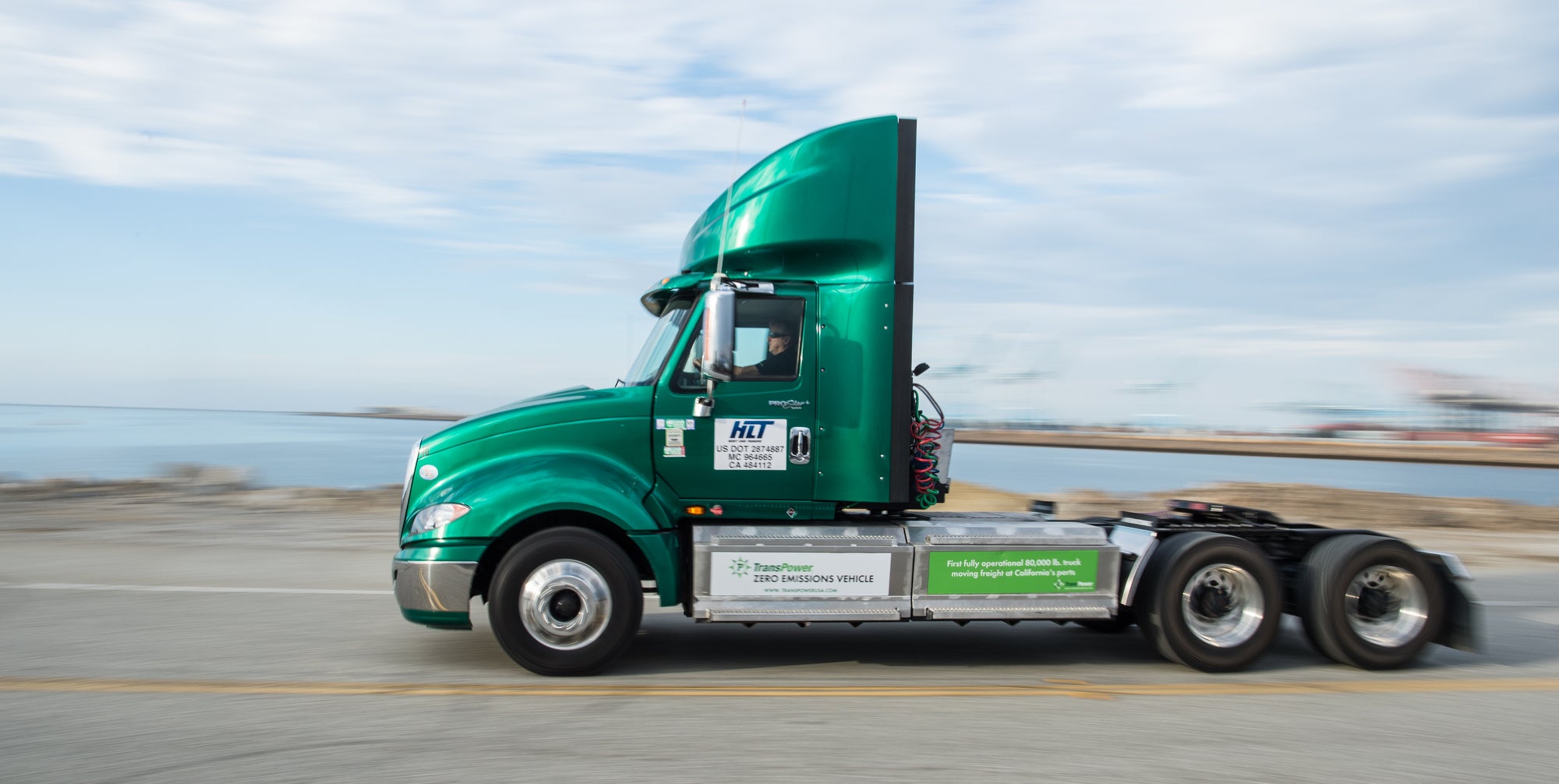 Green truck driving on road next to water.