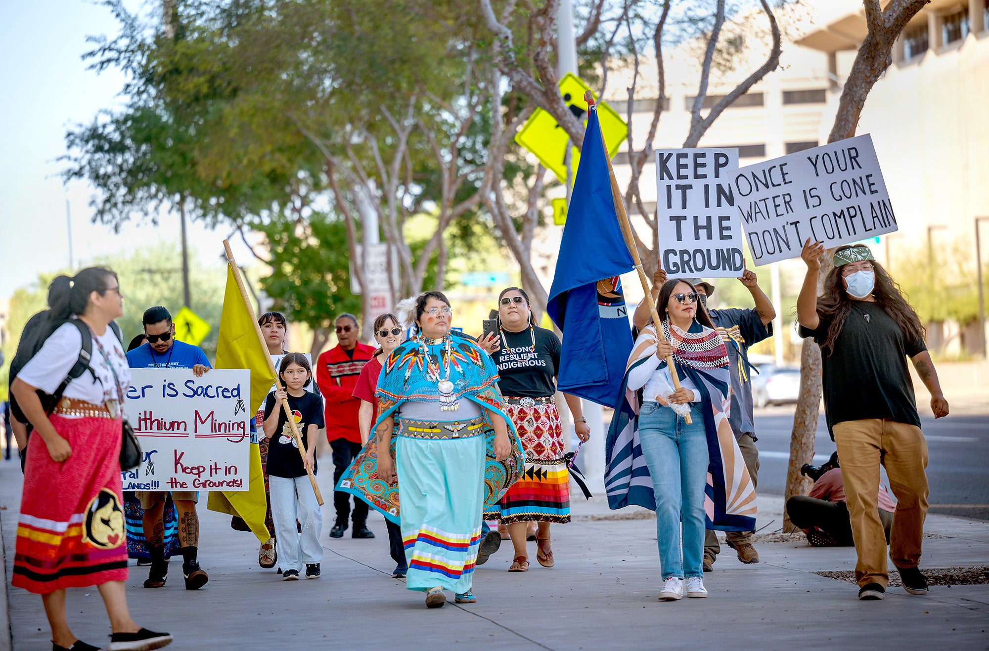 Tribal Rights Record A group of people march on a sidewalk holding flags and signs protesting mining.