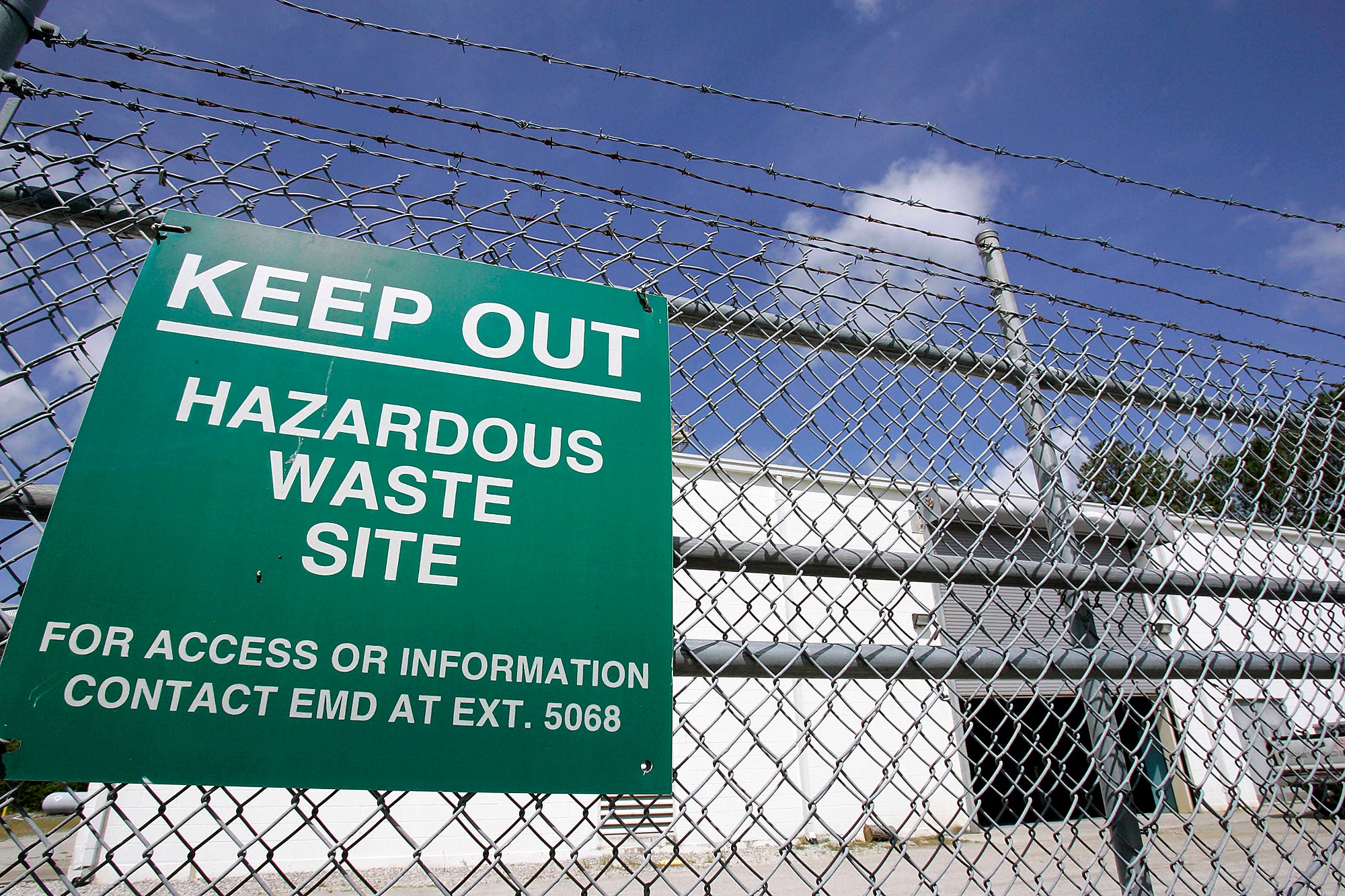 Toxic TCE Plume A green sign reading "Keep out hazardous waste site. For Access or information contact EMD at Ext. 5068" hangs on a chain link fence in front of a white cinder block building.