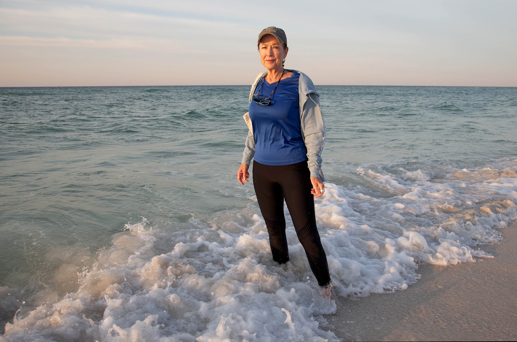 A woman wearing a hat, sweatshirt and black pants stands in ankle-deep water with the ocean behind her, looking directly at the camera with a serious expression.